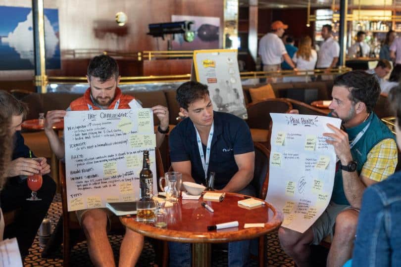 Three men sit in a row. The two on the outsides hold up large post-it boards with writing on them. The man in the middle reads the post-it board on the right.