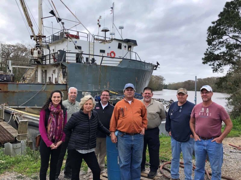A group of seven people stand together outdoors, in front of a large fishing boat parked on the banks of a river in Louisiana. They are looking at the camera and smiling.