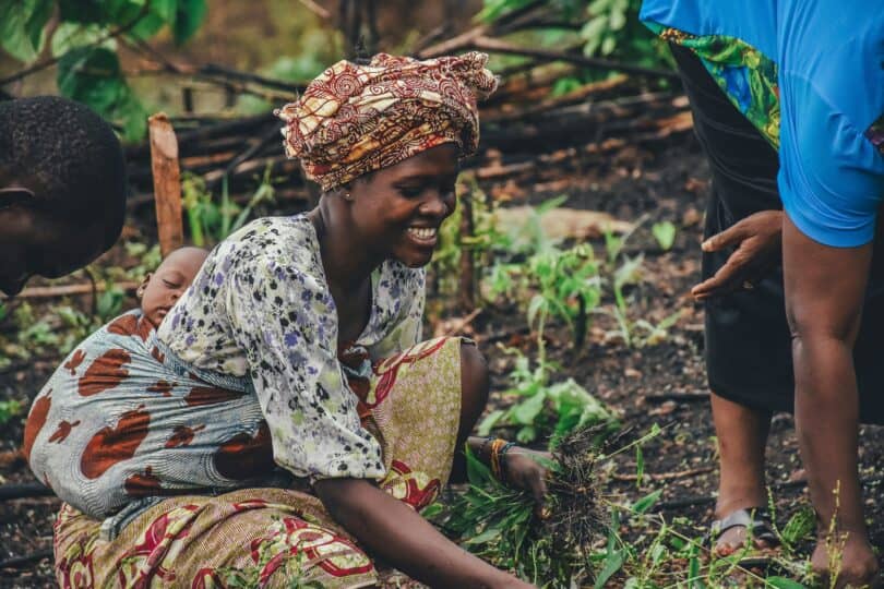 A smiling woman picks produce in a field. She has a small baby strapped to her back.
