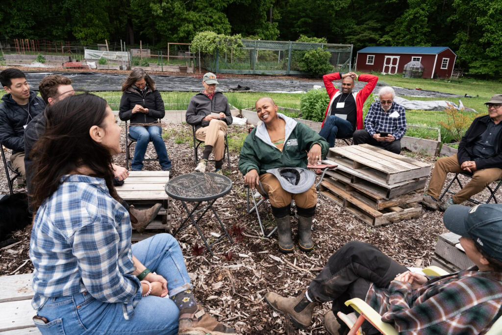 A group of people sit in a circle outside. They face a woman in the center of the circle who is smiling and talking.