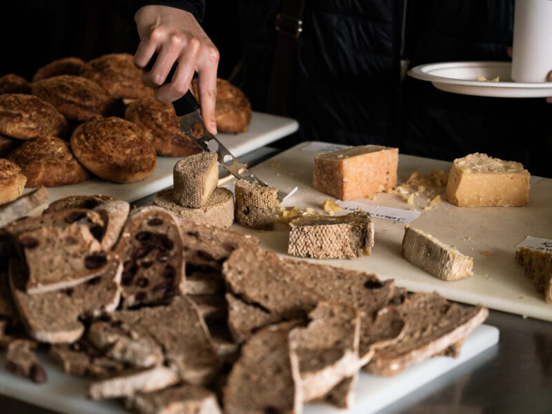 A woman's hand reaches in, cutting a block of cheese on a table covered by cheeses.